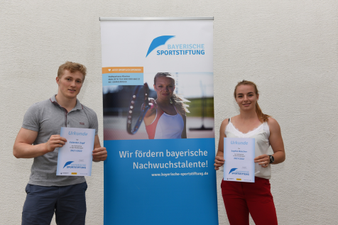 Mann und Frau stehen neben einen Aufsteller der Kuratoriumssitzung